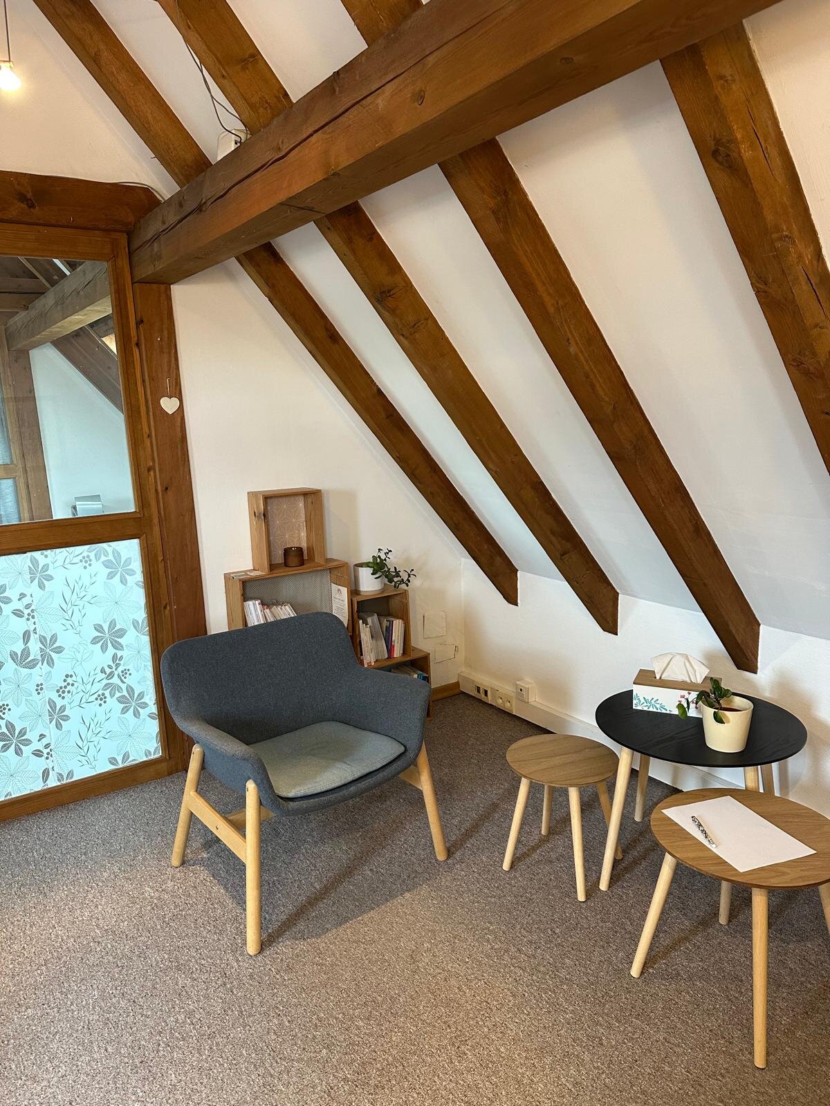 Salle d'attente du cabinet d'Aurélie Roethinger à Reichstett — verrière à motifs floraux, fauteuil et bibliothèque dans une ambiance chaleureuse aux poutres apparentes.